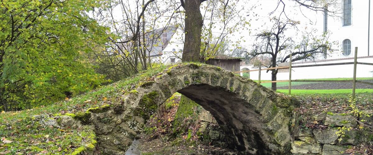 A stone bridge over a small stream in a park with trees and a building in the background.