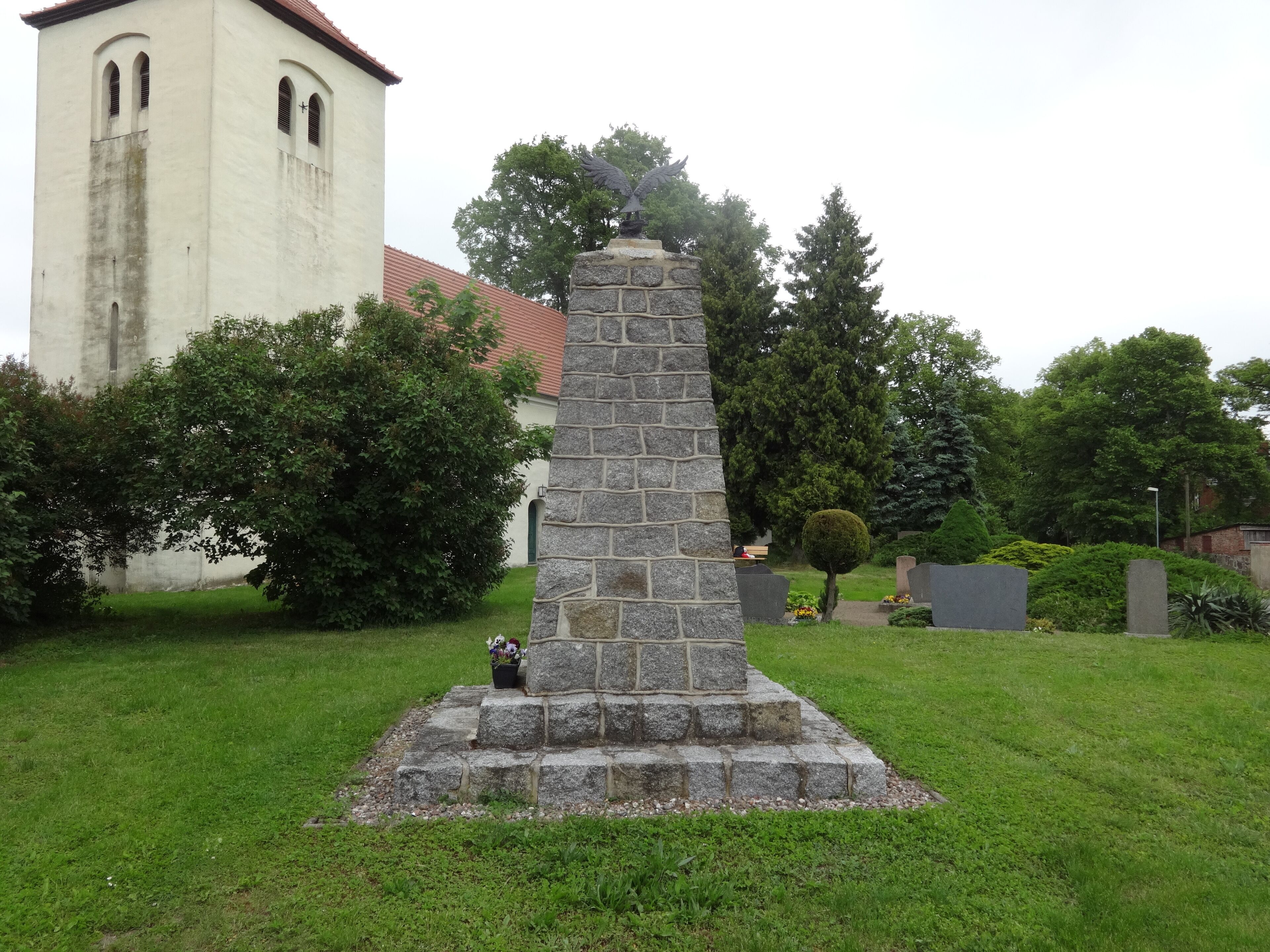 A stone monument with an eagle statue, surrounded by greenery and a church in the background.