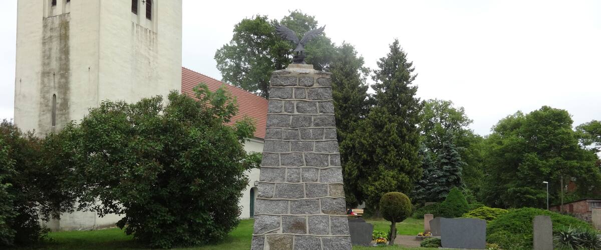A stone monument with an eagle statue, surrounded by greenery and a church in the background.