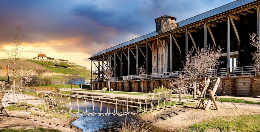 A large industrial building with a metal framework, a small water body in front, and a hilly landscape in the background.
