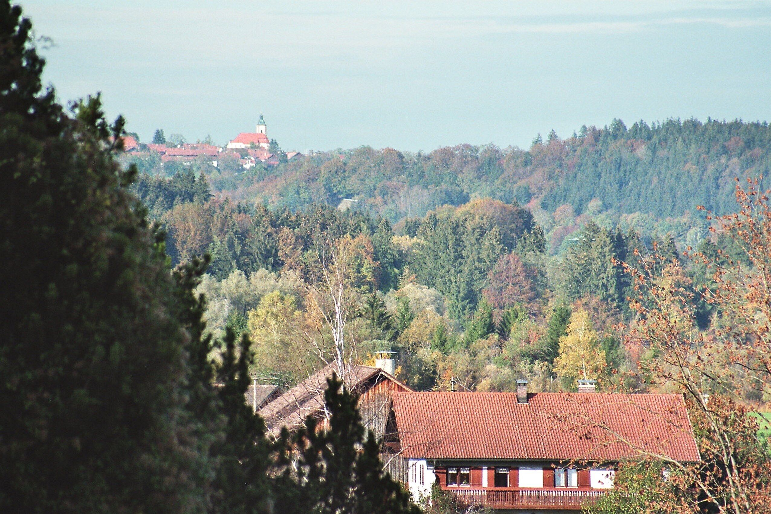 A rural landscape with a red-roofed house, a church with a green roof, and a forested hill in the background.