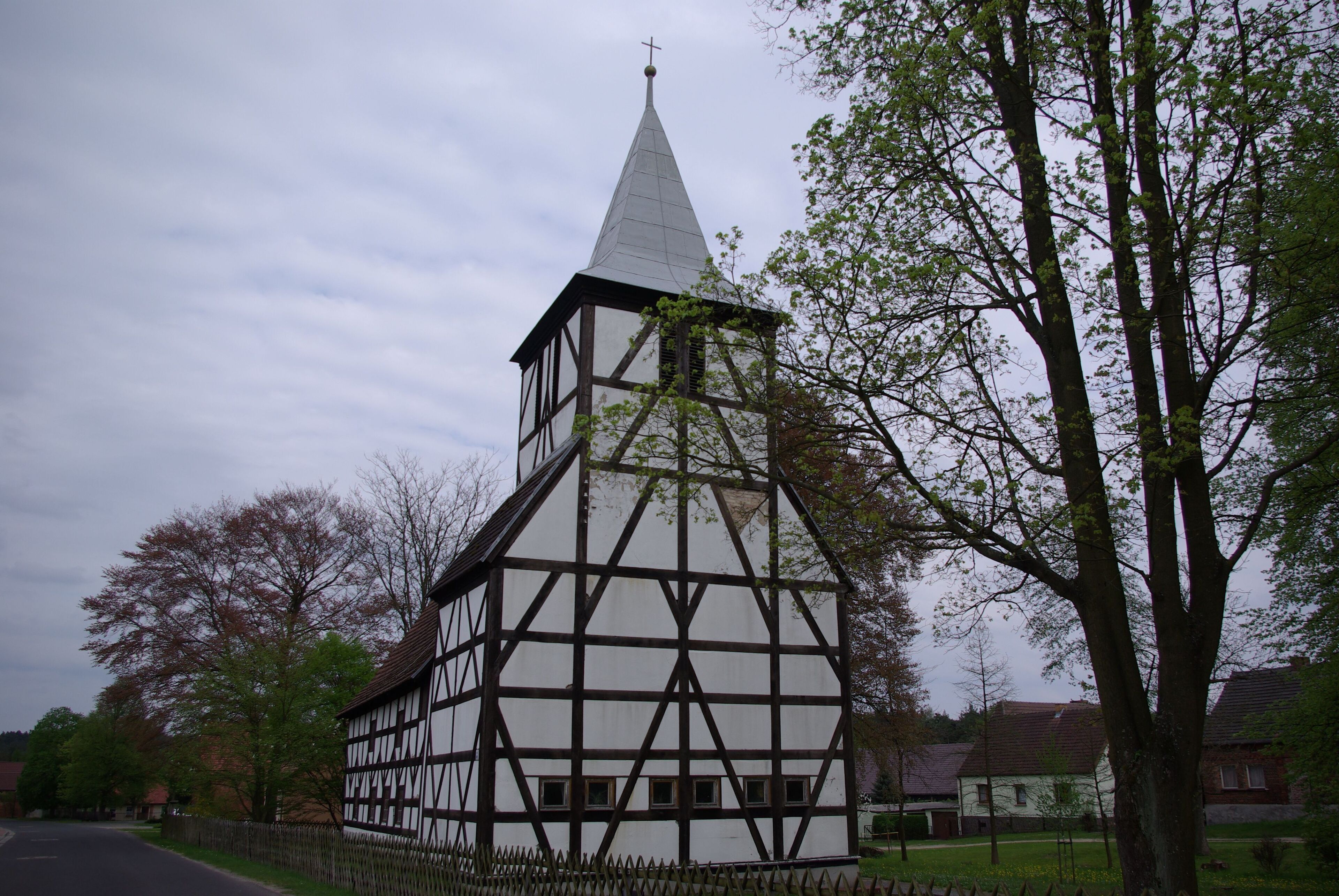 A half-timbered church with a pointed steeple and a cross on top, surrounded by trees and a fence.