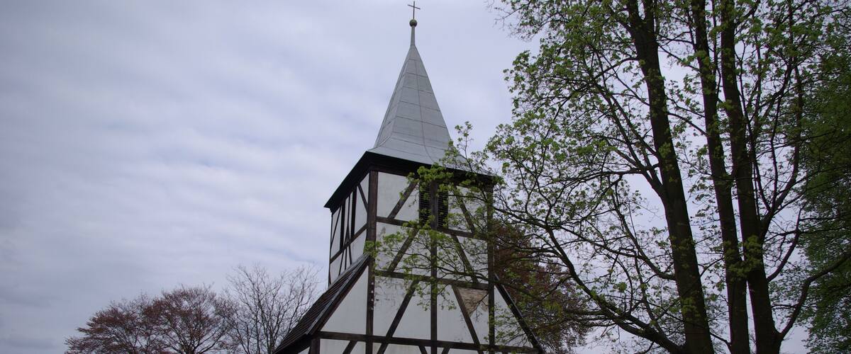 A half-timbered church with a pointed steeple and a cross on top, surrounded by trees and a fence.