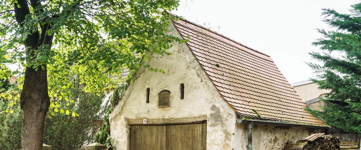 A rustic building with a wooden door and a tiled roof, surrounded by trees and a fence.