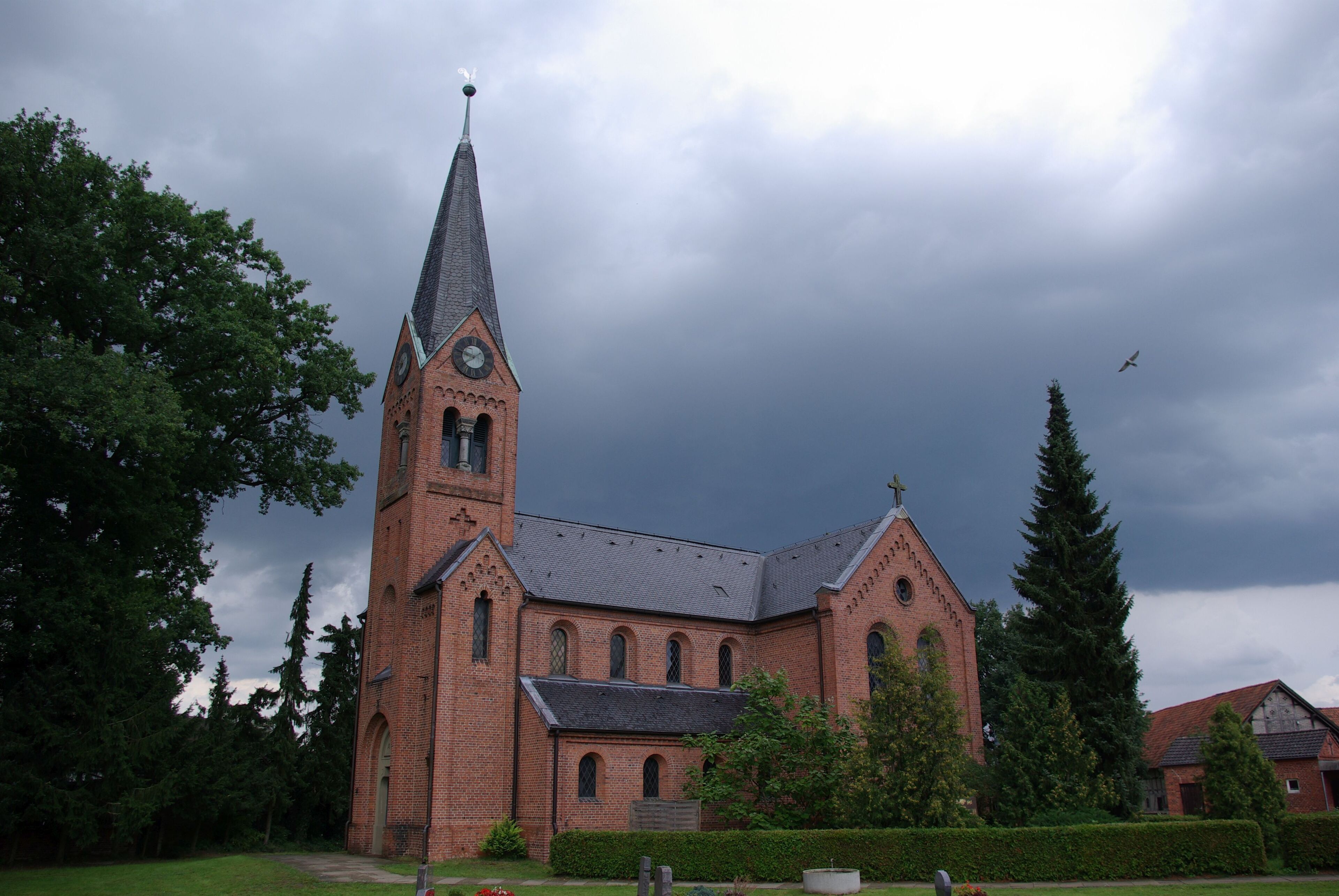 A brick church with a tall steeple and a clock, surrounded by trees and a hedge.