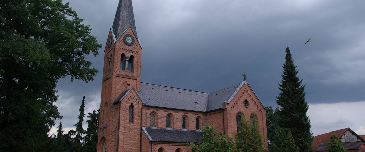 A brick church with a tall steeple and a clock, surrounded by trees and a hedge.