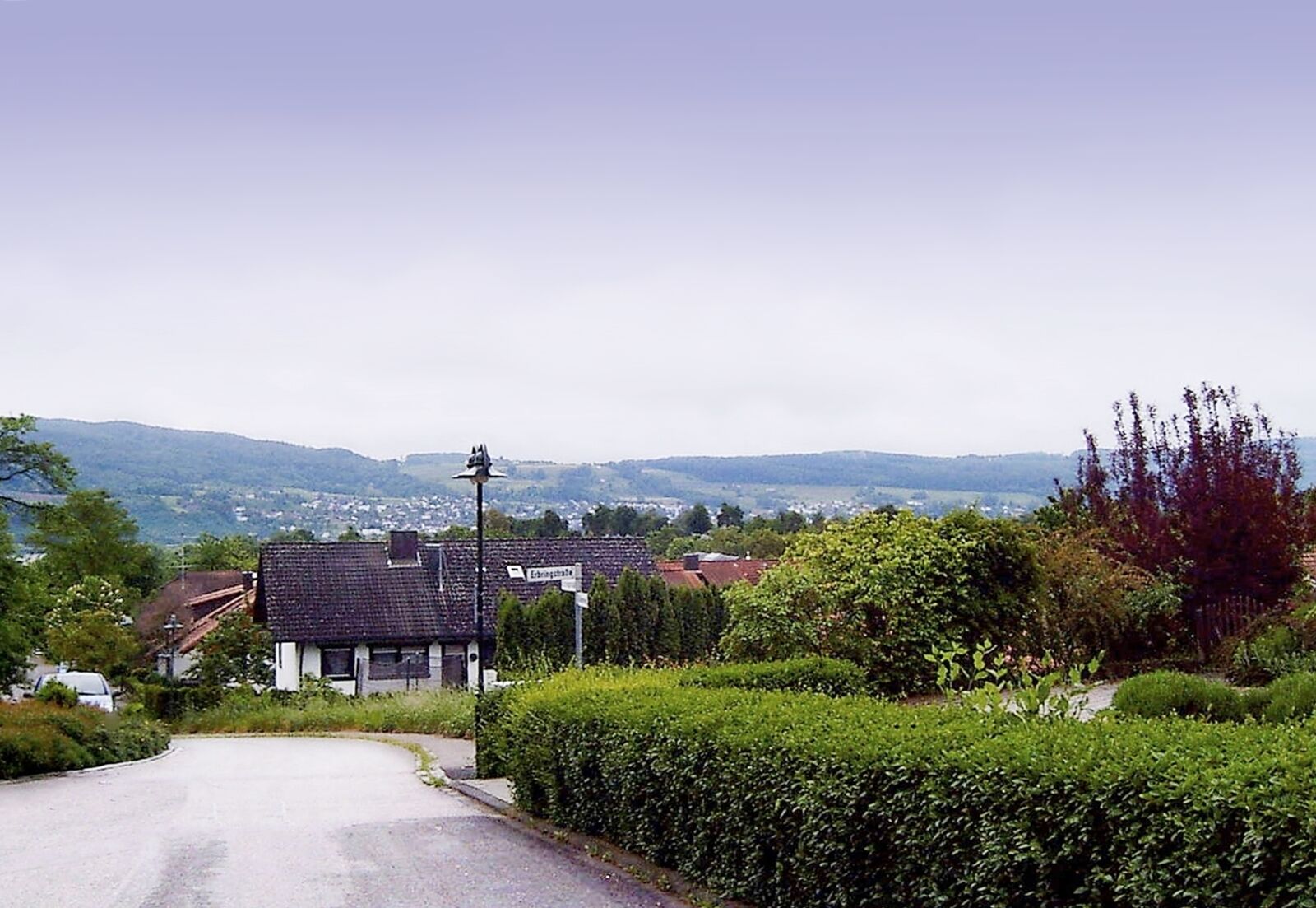A rural road with a house, hedges, and a distant view of hills and a town.
