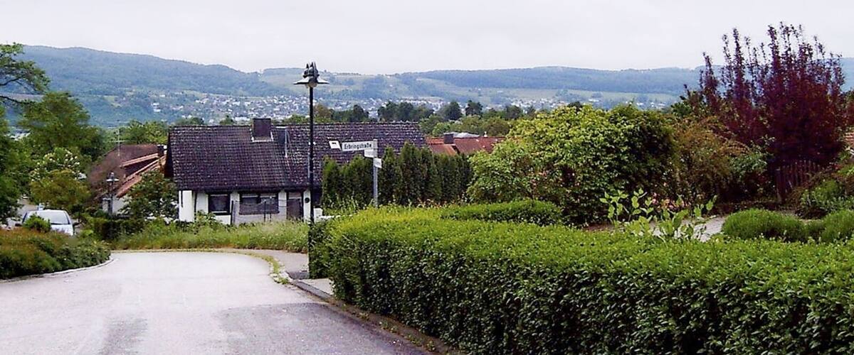 A rural road with a house, hedges, and a distant view of hills and a town.