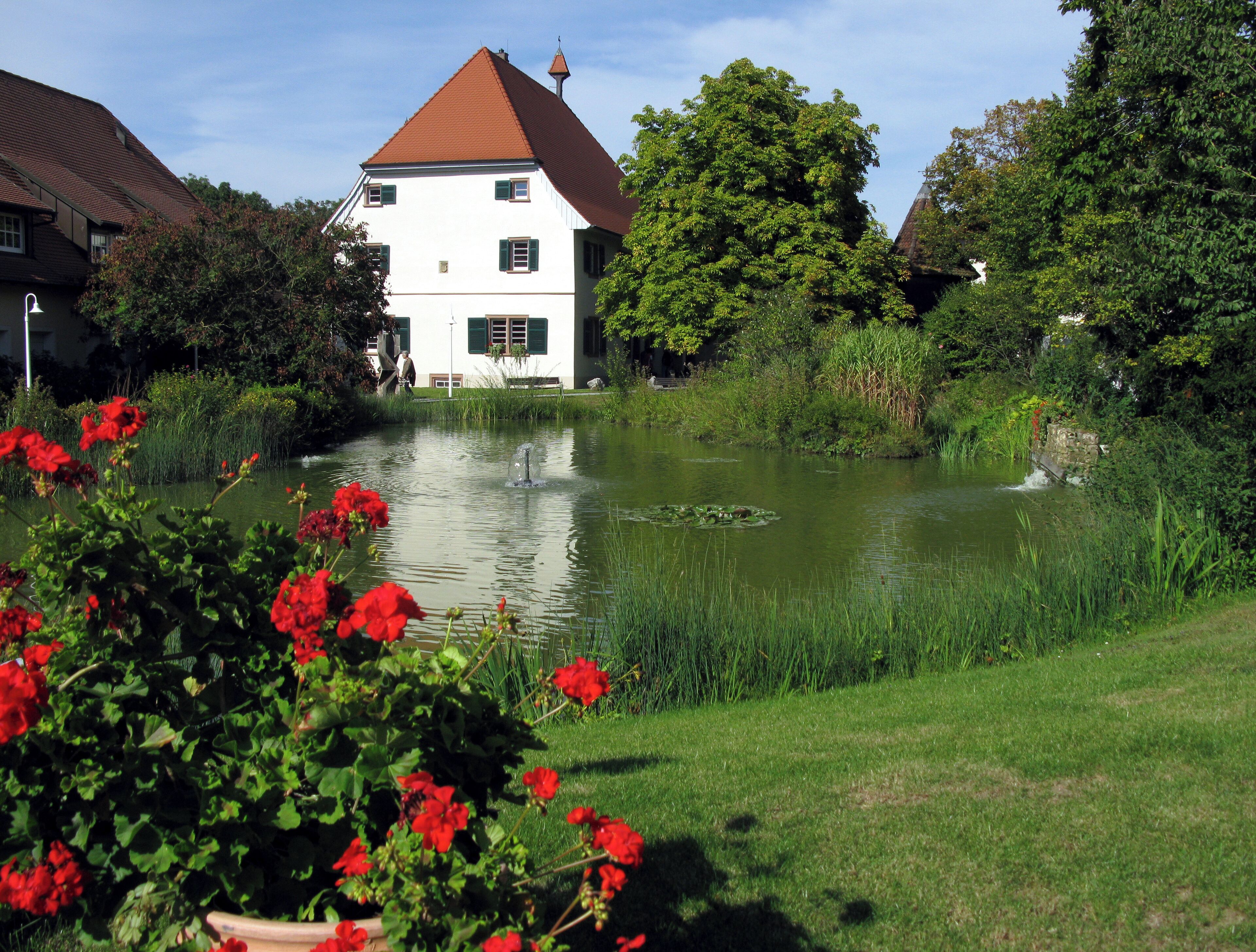 A pond with a fountain, surrounded by lush greenery and a white building with a red roof.