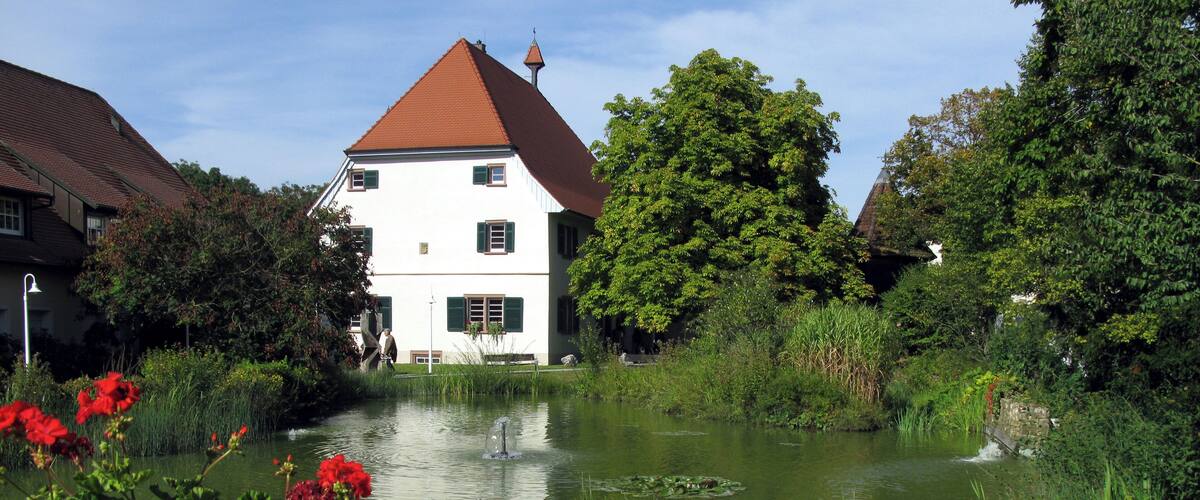A pond with a fountain, surrounded by lush greenery and a white building with a red roof.