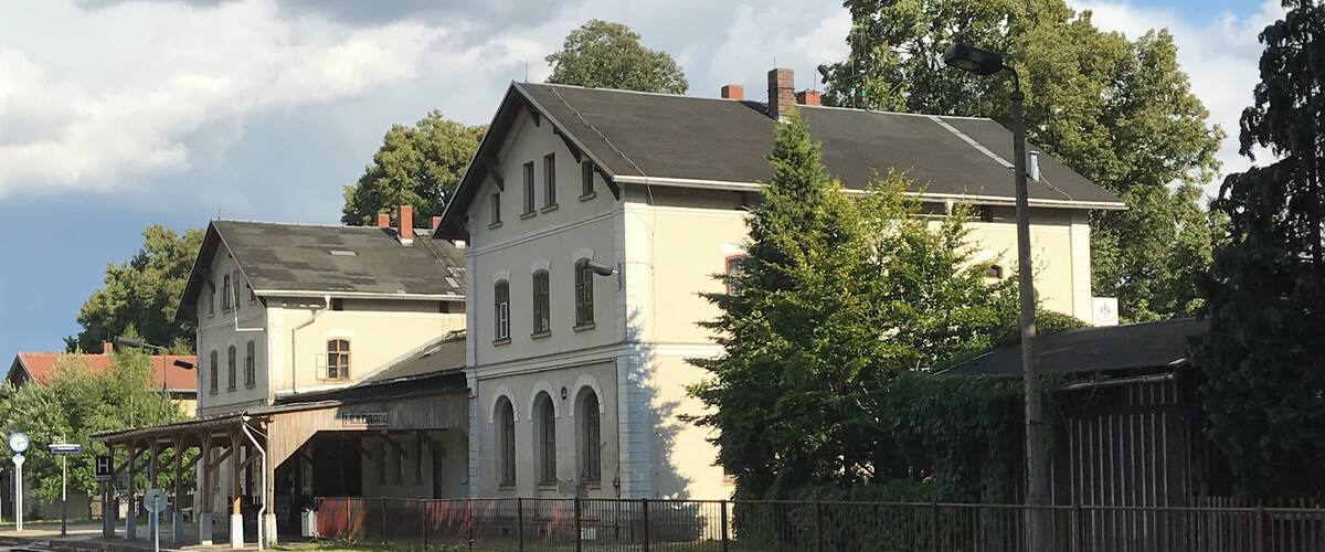 A train station with a white building, tracks, and trees.