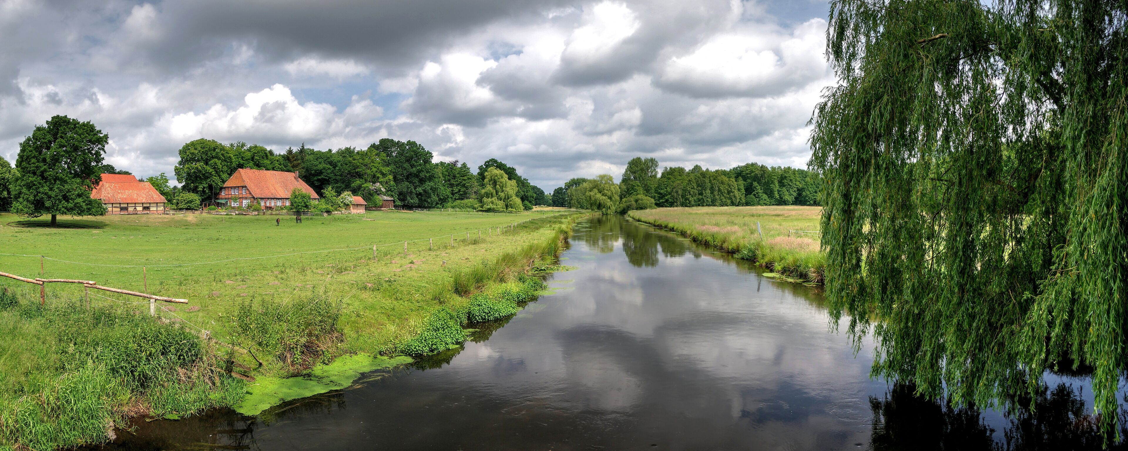 A calm river with a wooden bridge, a red-roofed house, and lush greenery.