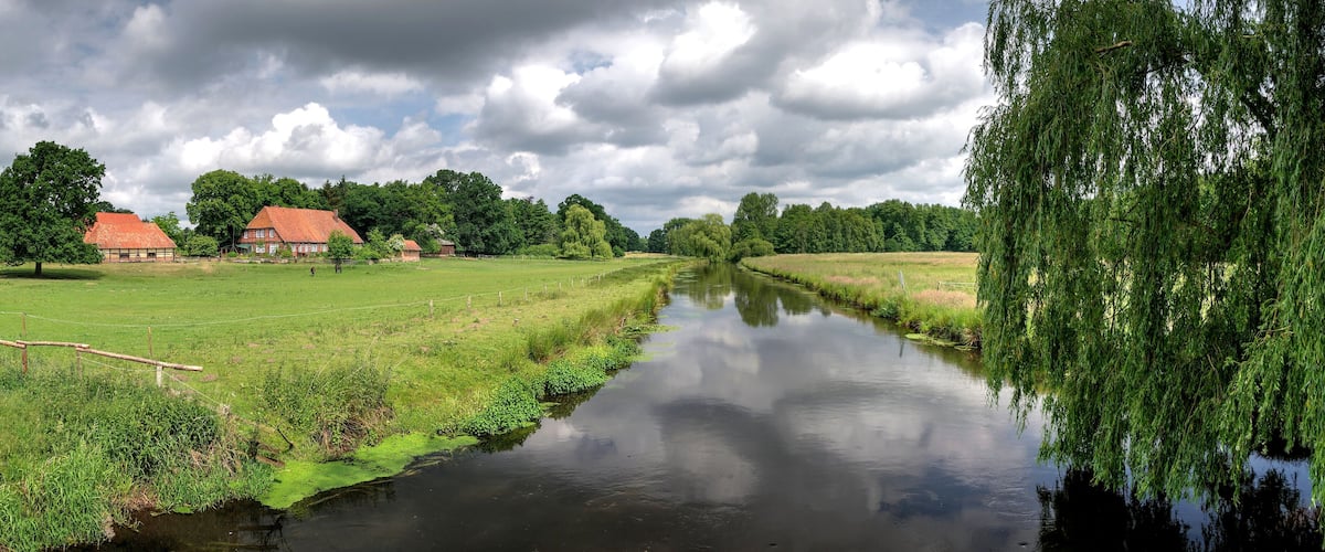 A calm river with a wooden bridge, a red-roofed house, and lush greenery.