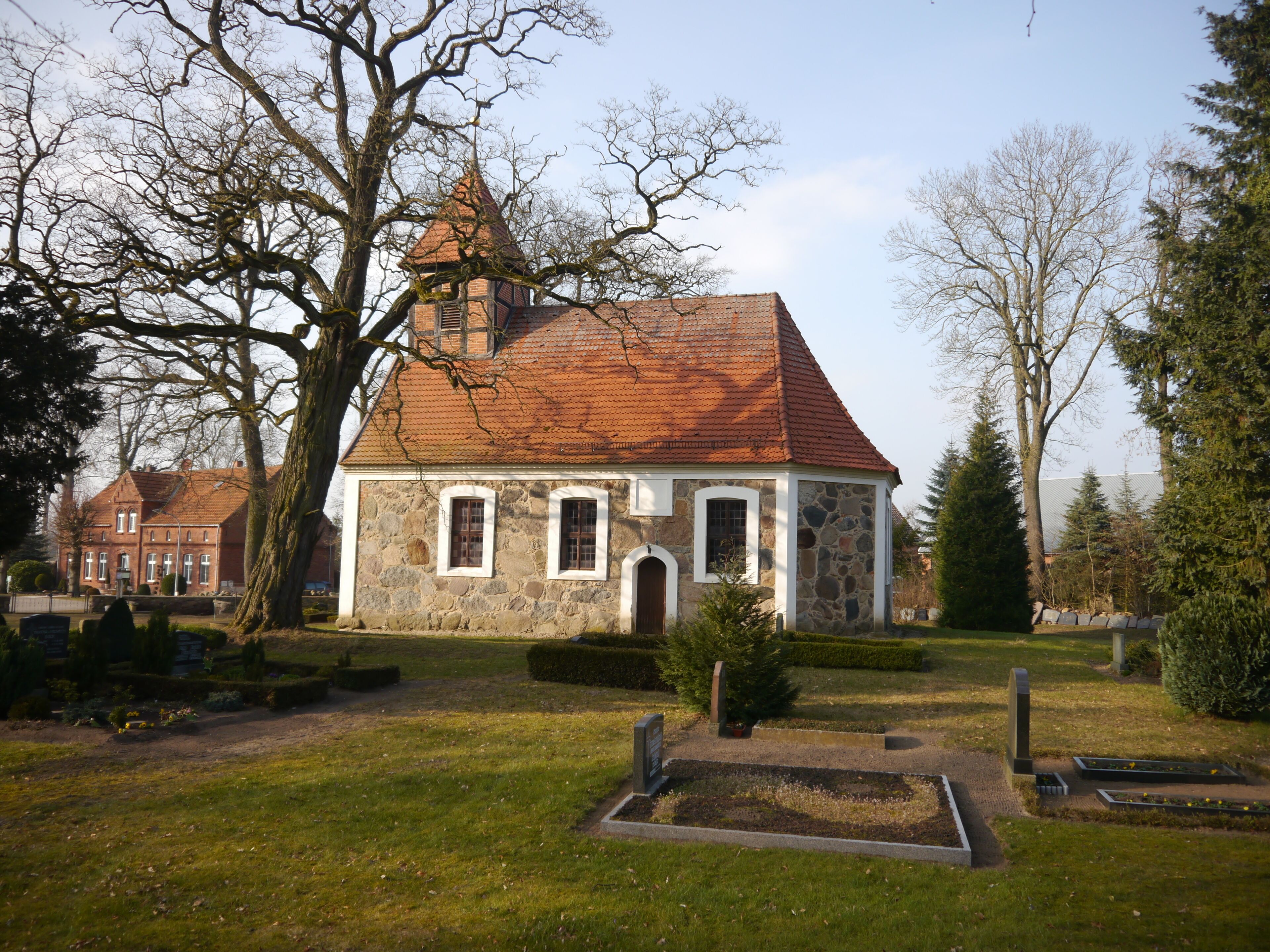A stone church with a red-tiled roof and a bell tower, surrounded by trees and a well-maintained lawn.