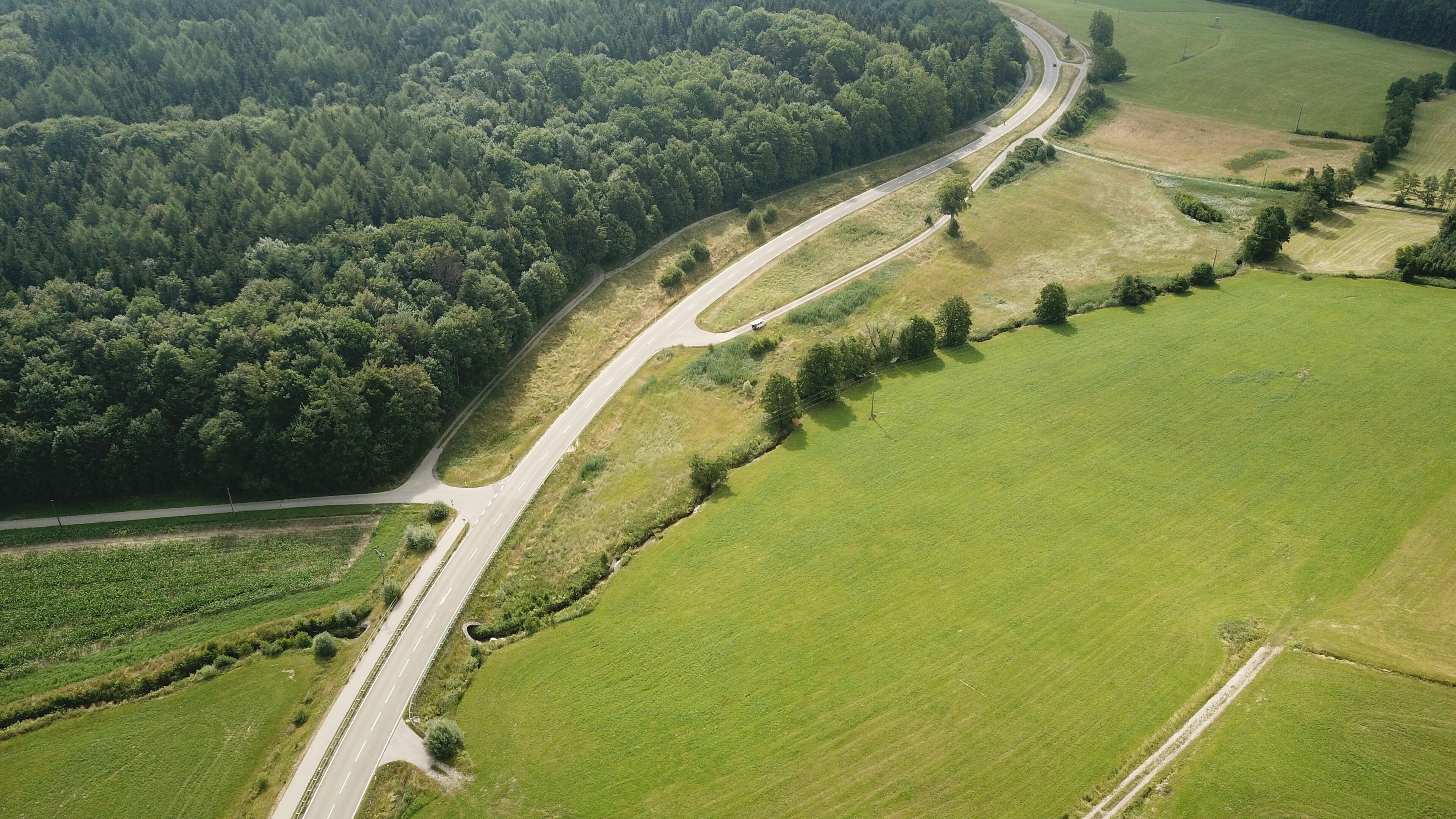 An aerial view of a winding road through a green landscape with fields and a dense forest.