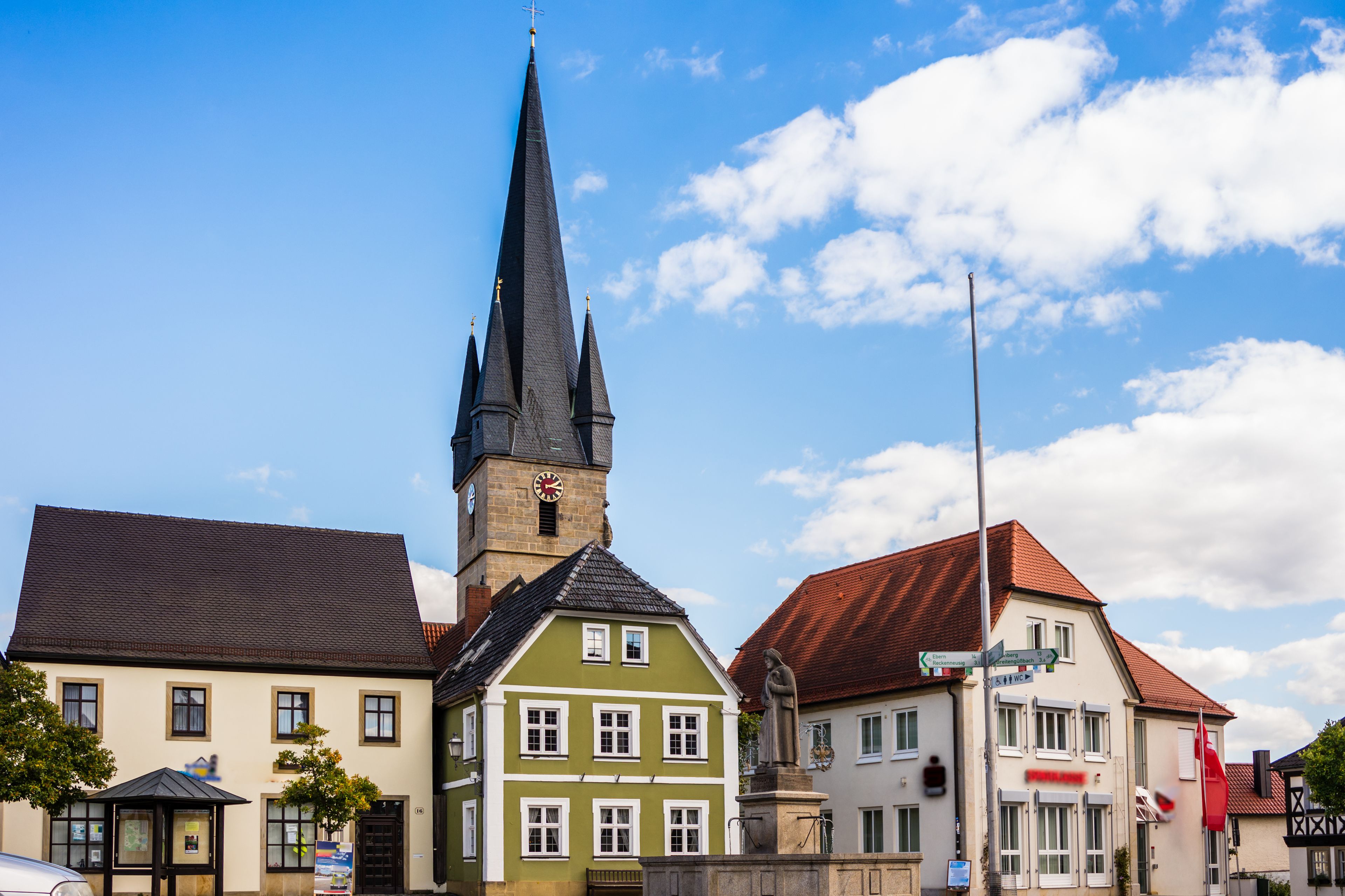En kirke med et højt spir, en grøn bygning med hvide vinduesrammer og en bygning med rødt tag og et skilt.