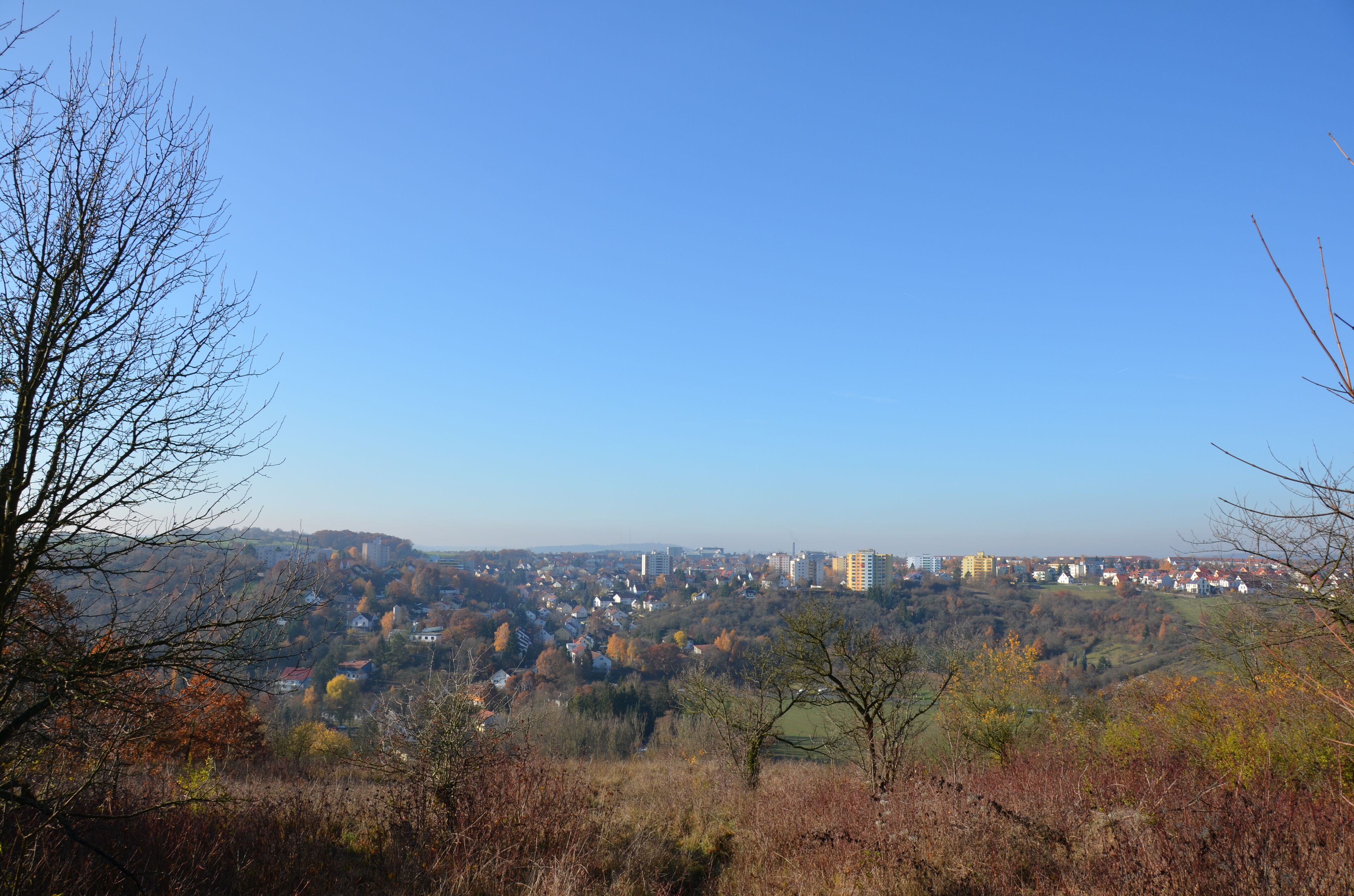 A hilly landscape with autumn-colored trees, a town in the valley, and a clear blue sky.