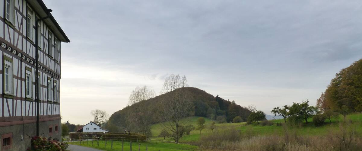 A rural road curving through a green landscape with a hill in the distance and a half-timbered building on the left.