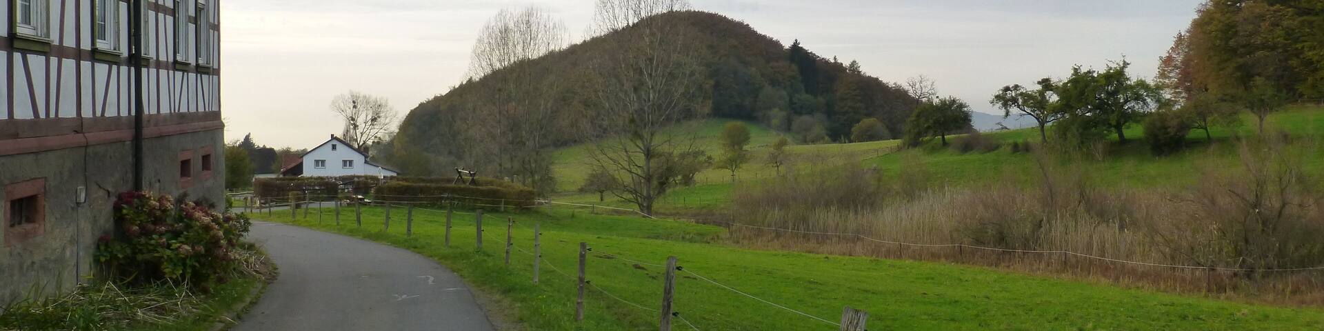 A rural road curving through a green landscape with a hill in the distance and a half-timbered building on the left.