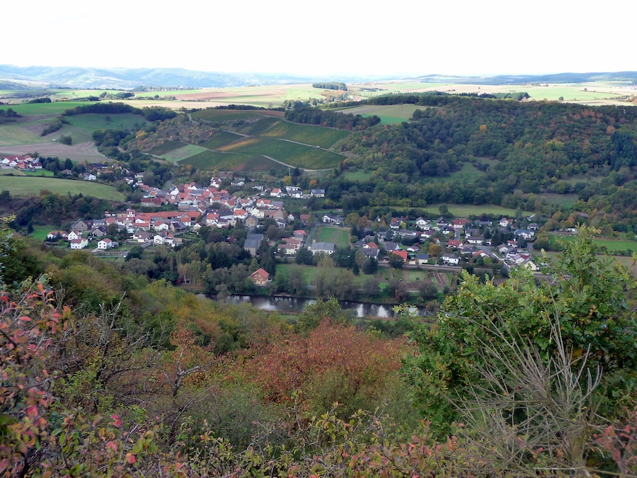 Ein Dorf eingebettet in ein Tal, mit einem Fluss, umgeben von sanften Hügeln und Feldern.