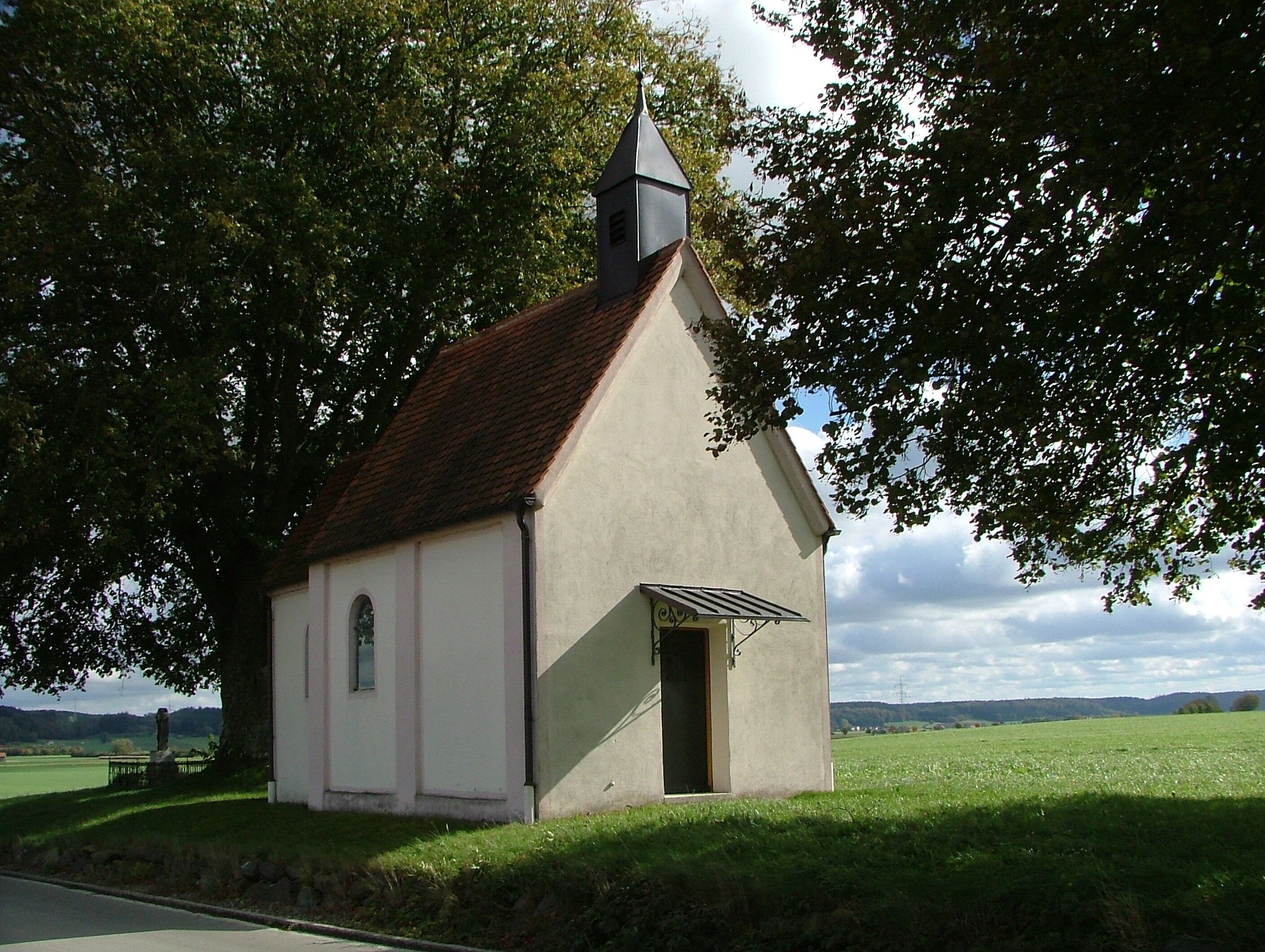 A small church with a steep roof and a bell tower, surrounded by lush green fields and trees.