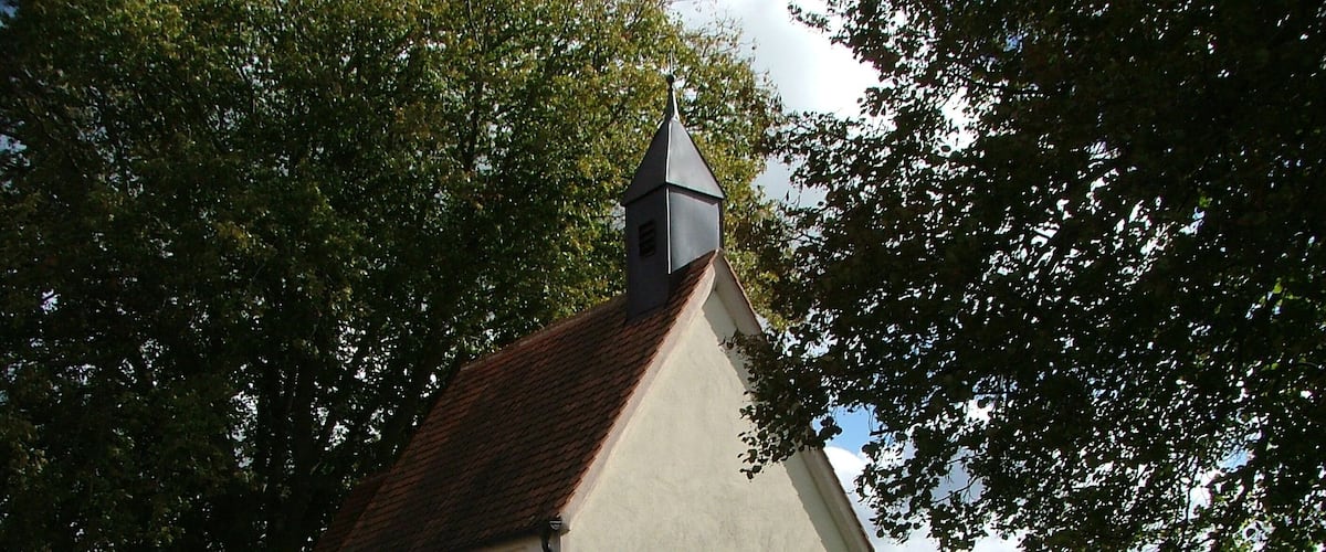 A small church with a steep roof and a bell tower, surrounded by lush green fields and trees.