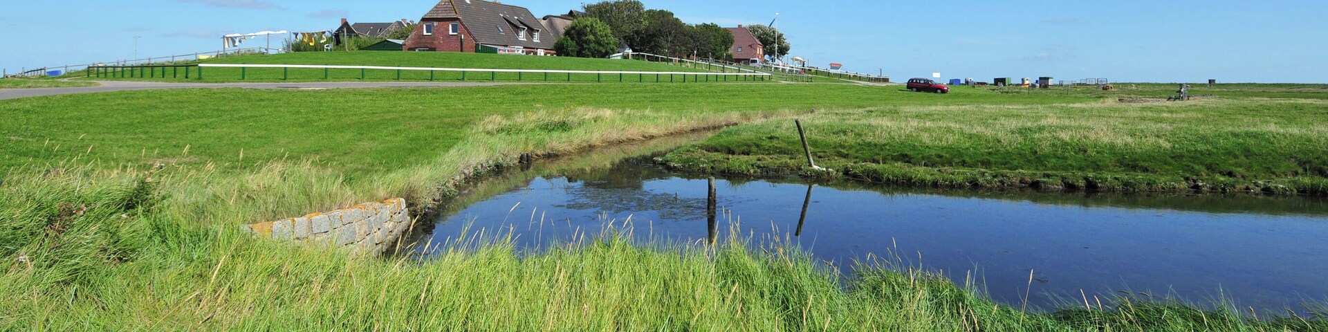 View to the Backenswarft on Hallig Hooge. The Hallig Hooge is the second largest of the ten islets in the Schleswig-Holstein Wadden Sea. Hallig Hooge is surrounded by an approximately 1.20 meters high stone dyke and is thus protected from light storm surges. Nevertheless Hallig Hooge is flooded completely two to five times a year. The Houses are built on high mounds. Therefore they can withstand the storm surges. The Hallig Hooge has twelve mounds. The Backenswarft is the second largest mound with a dozen buildings. The buildings surround the pond with rainwater.