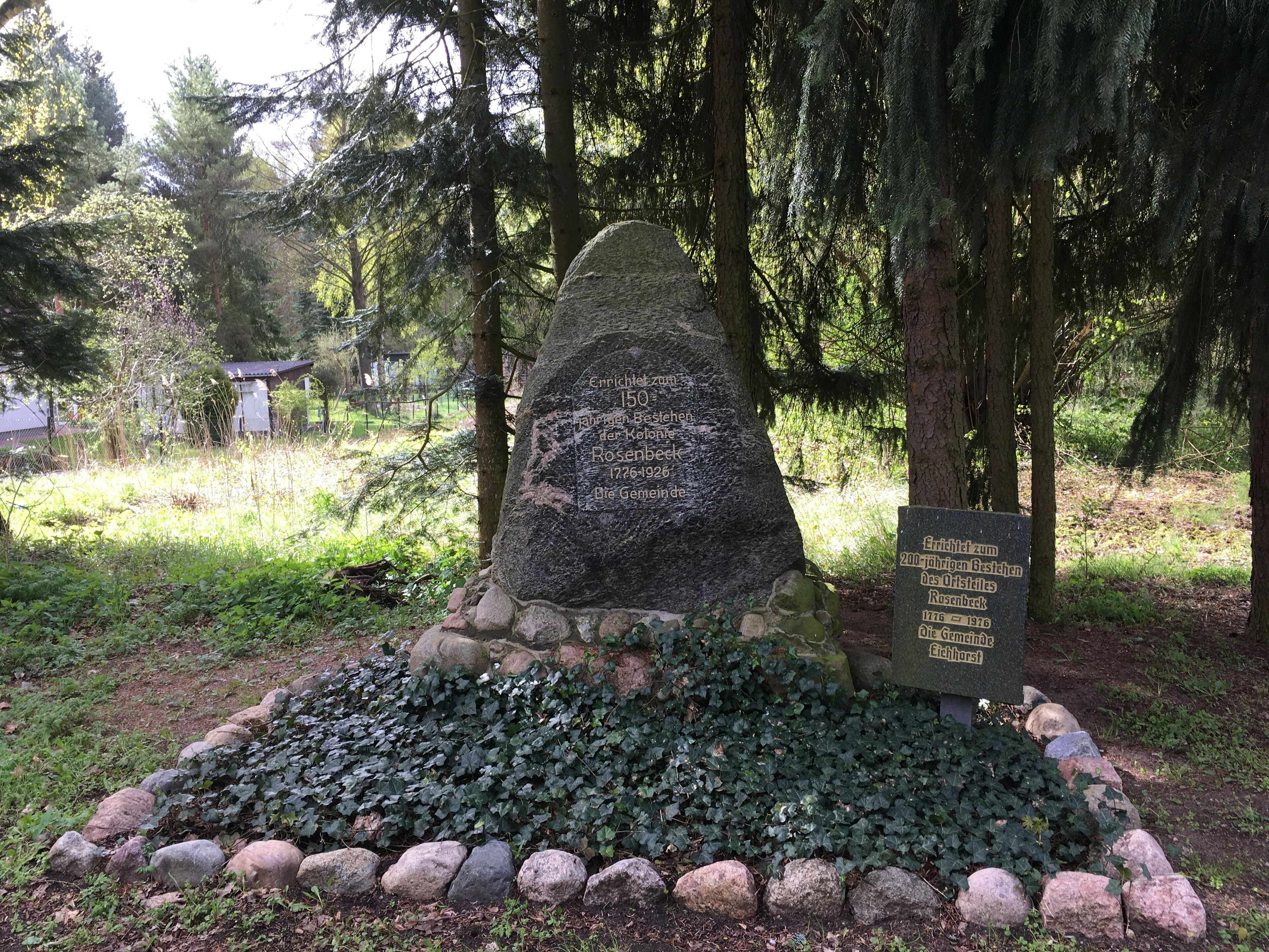 A memorial stone with inscriptions, surrounded by a stone border and greenery.