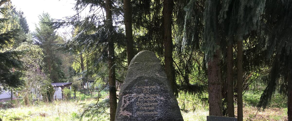 A memorial stone with inscriptions, surrounded by a stone border and greenery.
