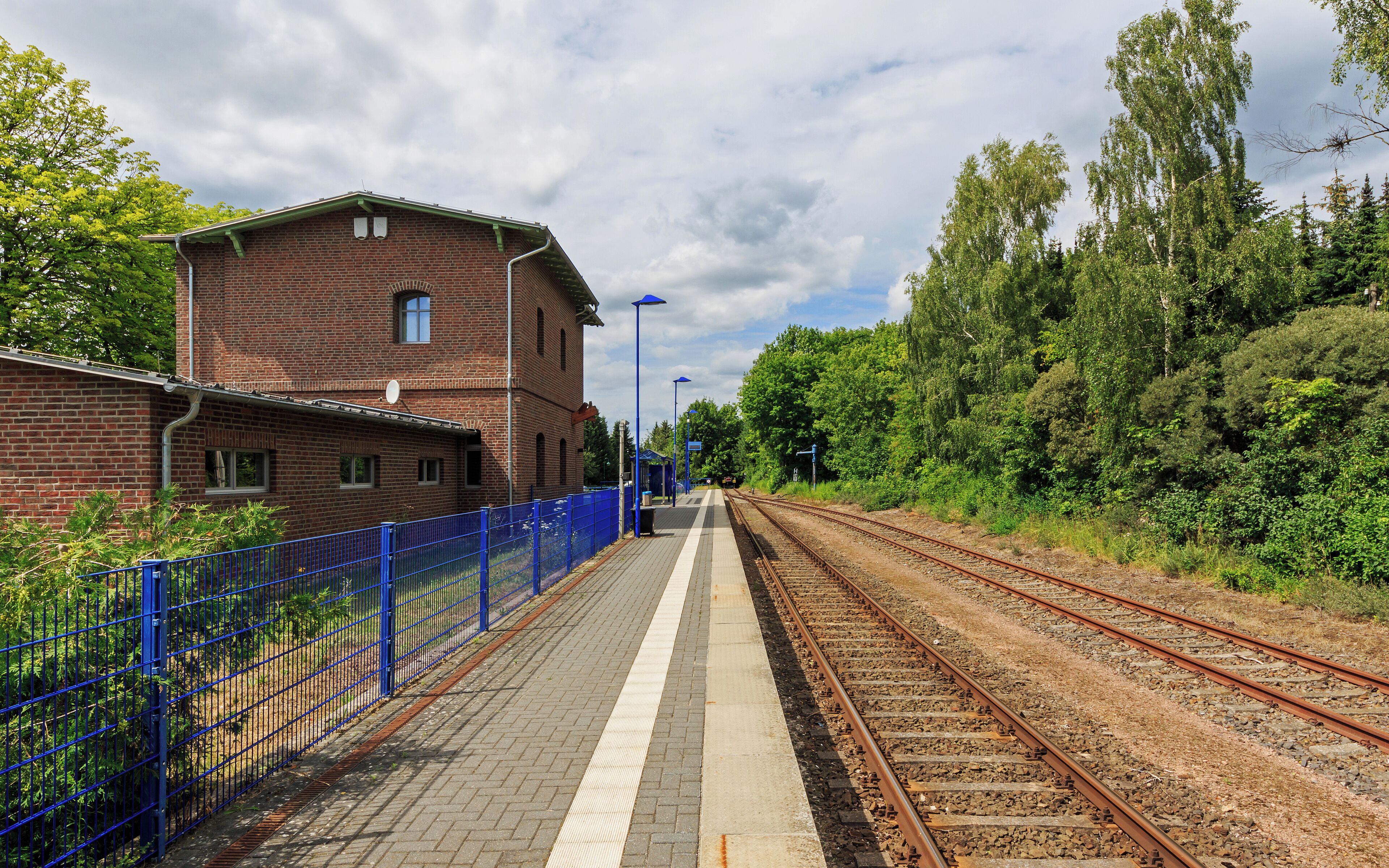 A train station platform with a brick building, blue fencing, and railway tracks.