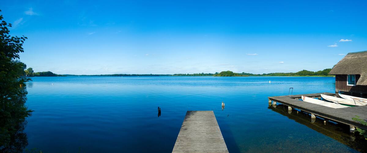 A calm lake with a wooden dock extending into the water, surrounded by greenery and a thatched-roof structure.