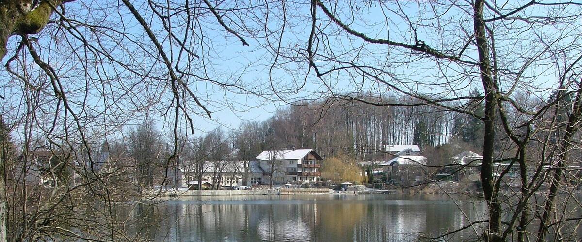 A lake with buildings in the background, surrounded by leafless trees and snow on the ground.