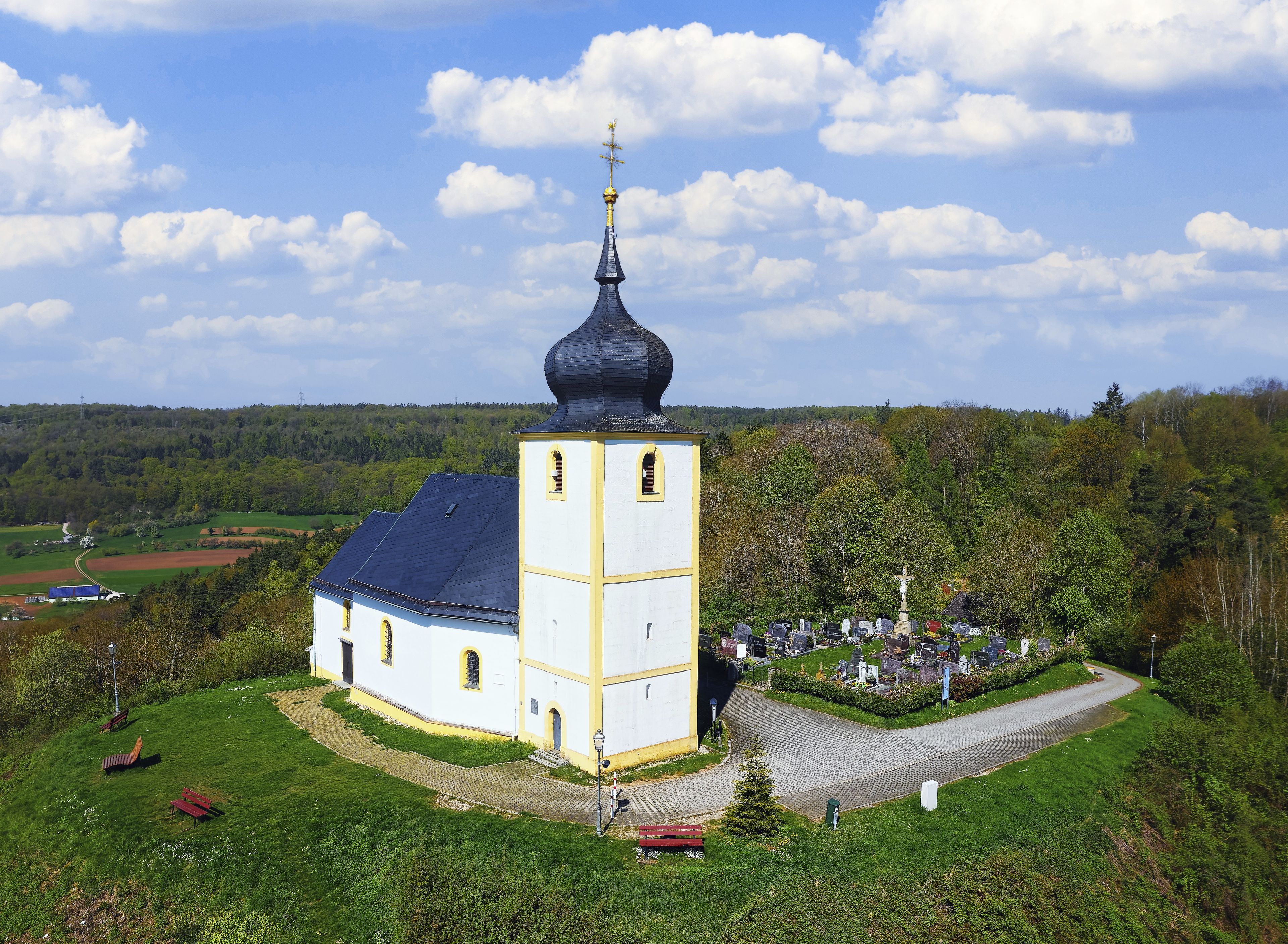 Eine weiße Kirche mit blauem Dach und schwarzer Kuppel, umgeben von einem Friedhof mit Grabsteinen und einem Kreuz.
