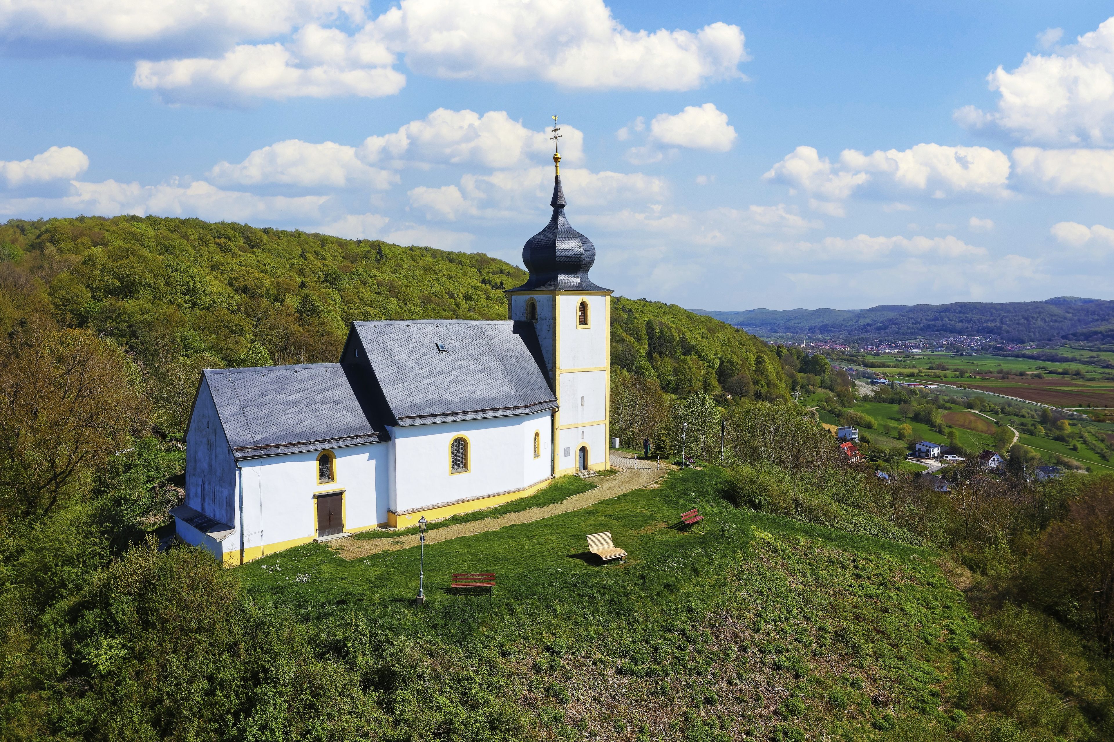 Eine weiße Kirche mit einem schwarzen Kirchturm auf einem Hügel, umgeben von Grün und einem Dorf in der Ferne.