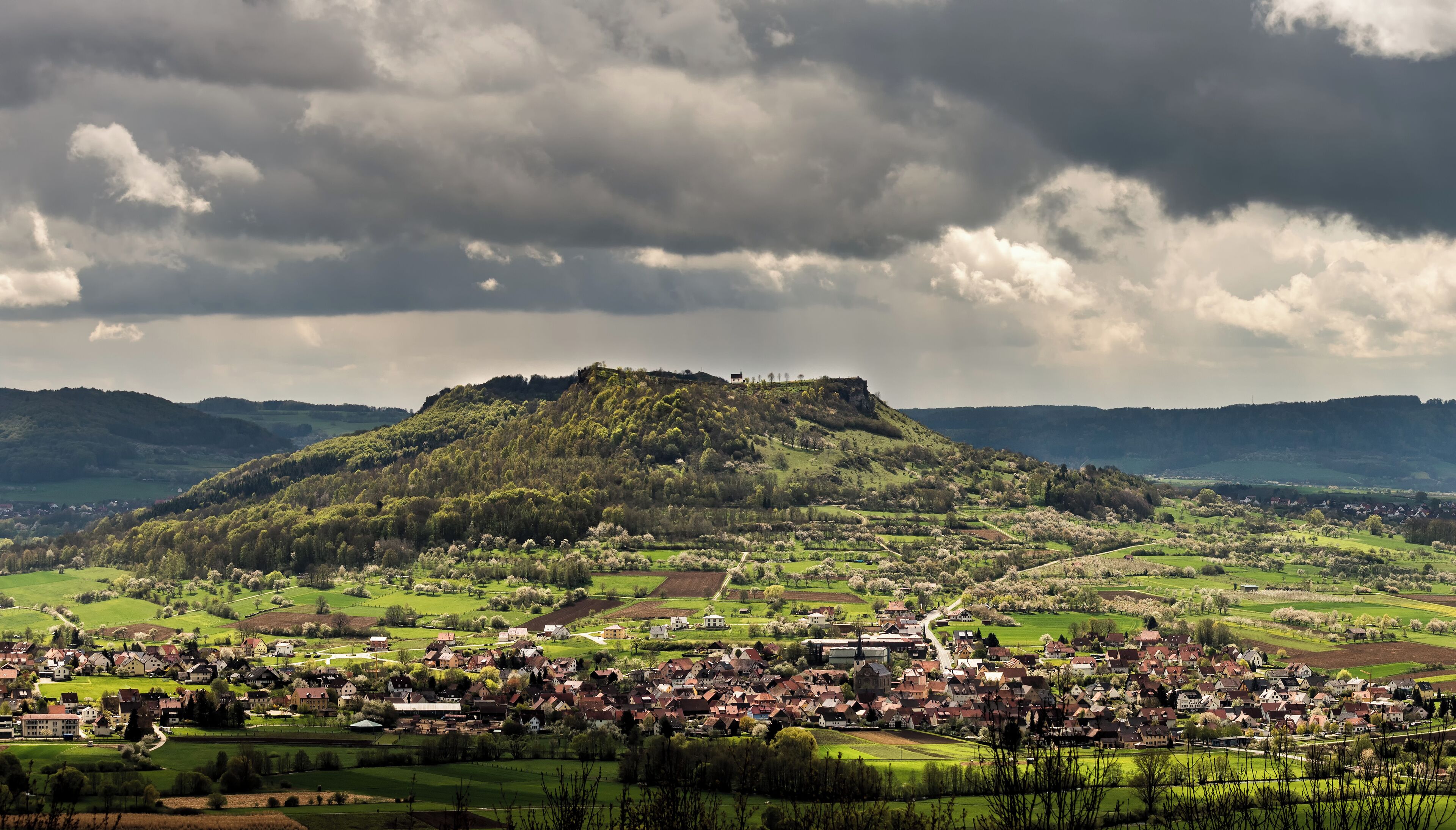 A village with a hilltop castle, surrounded by green fields and a cloudy sky.