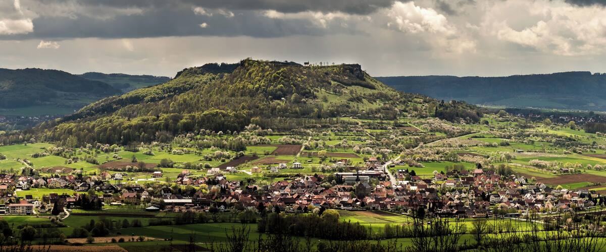 A village with a hilltop castle, surrounded by green fields and a cloudy sky.