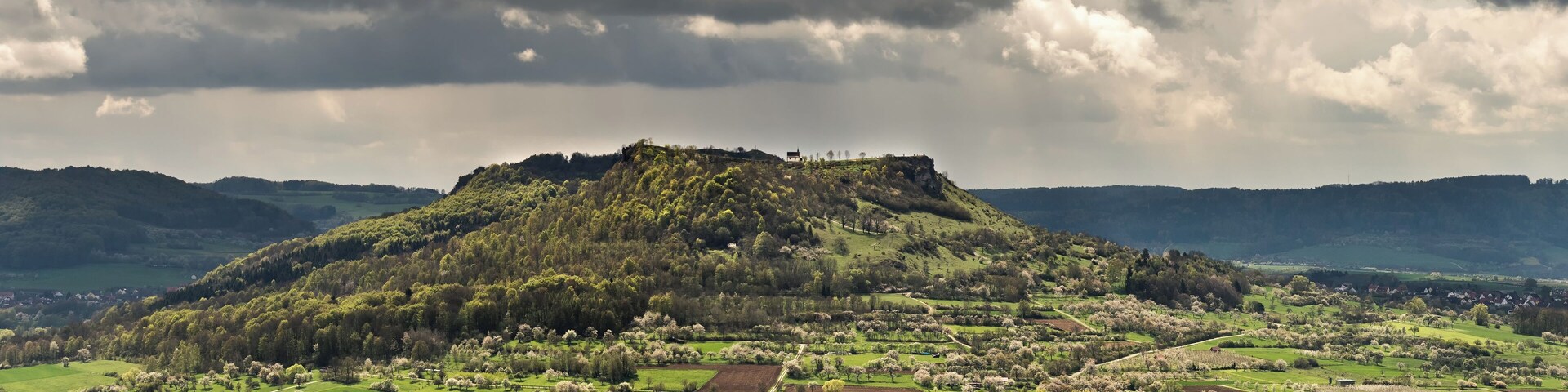 A village with a hilltop castle, surrounded by green fields and a cloudy sky.
