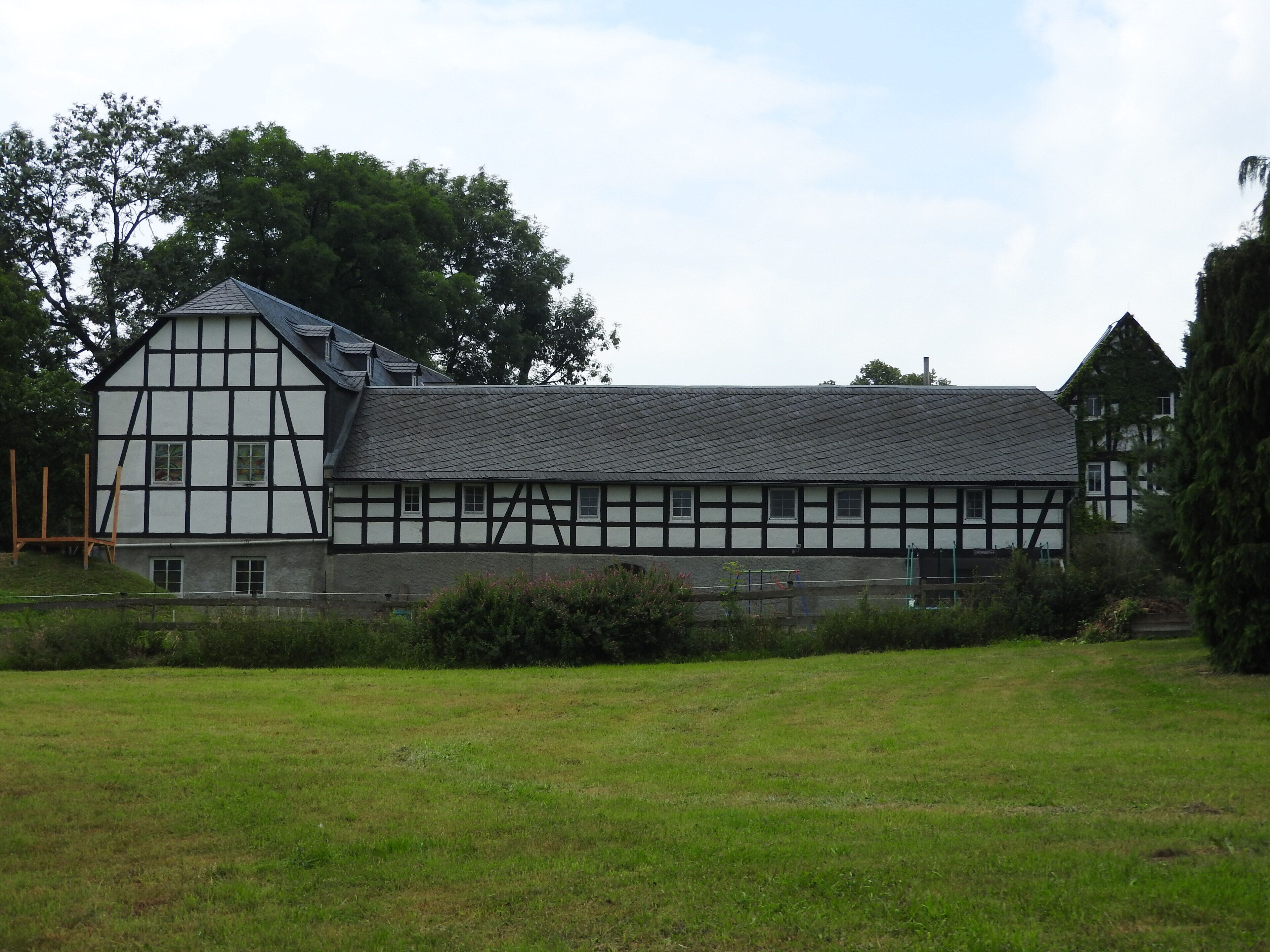 A traditional half-timbered house with a sloped roof, surrounded by a grassy field and trees.