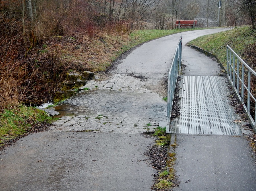 A paved path curving through a wooded area with a metal bridge over a small stream.