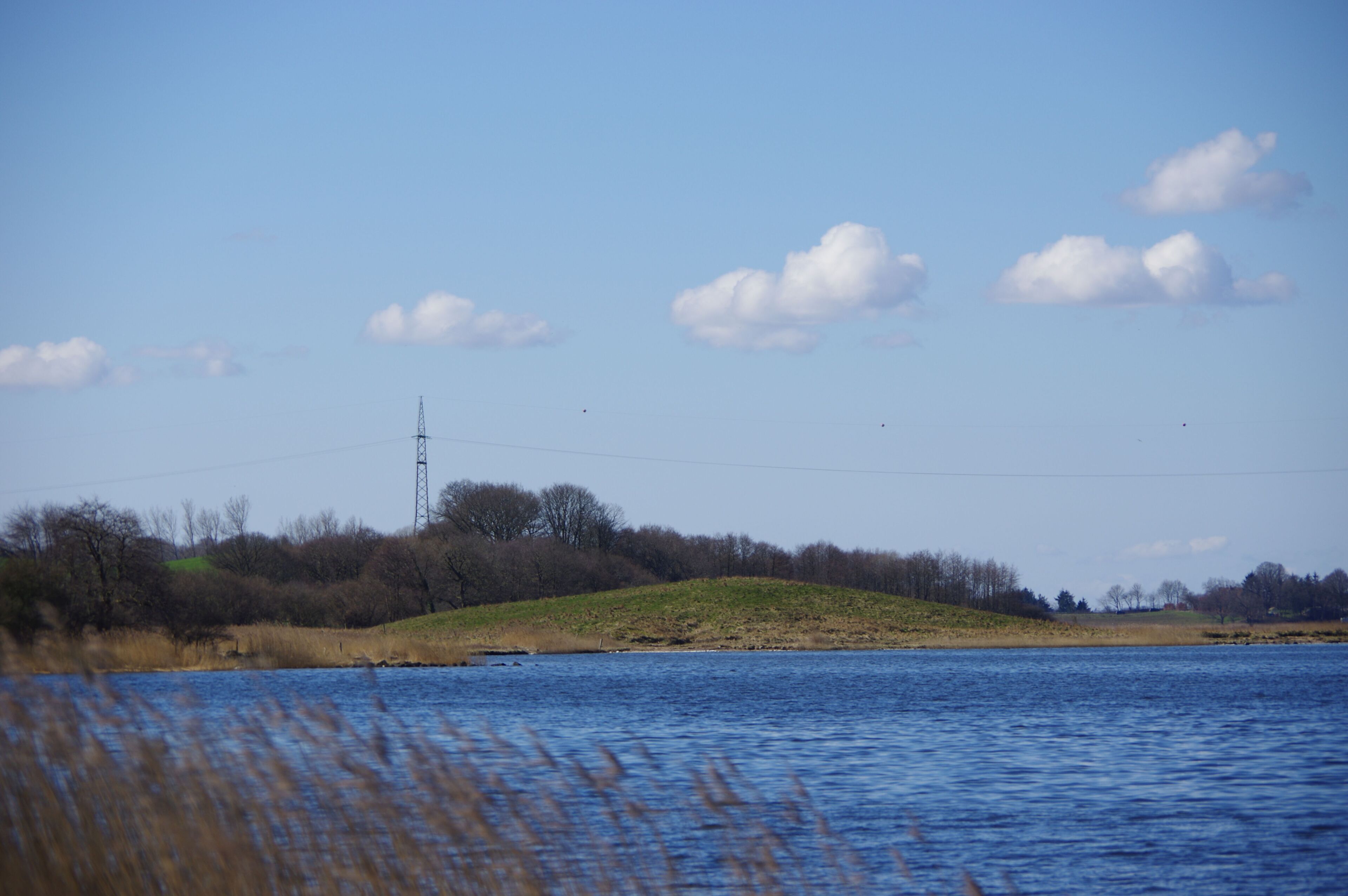 A calm lake with a grassy hill and a power line tower in the distance.