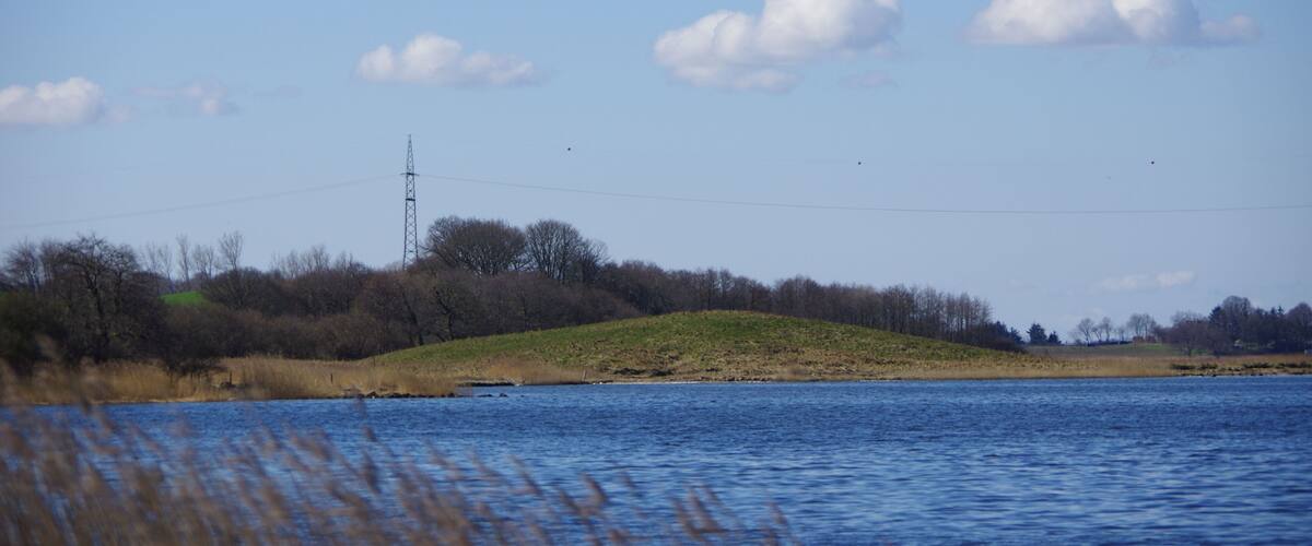 A calm lake with a grassy hill and a power line tower in the distance.