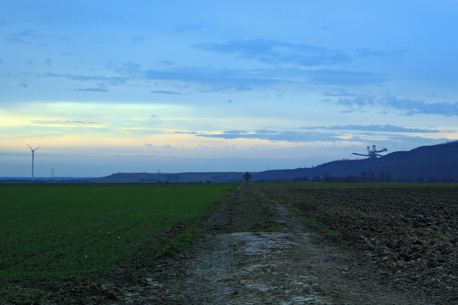 Un paysage rural avec un chemin de terre qui traverse des champs, une éolienne et un hélicoptère visible au loin.