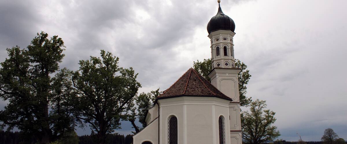 A white church with a black dome and a cross on top, surrounded by trees and a cloudy sky.