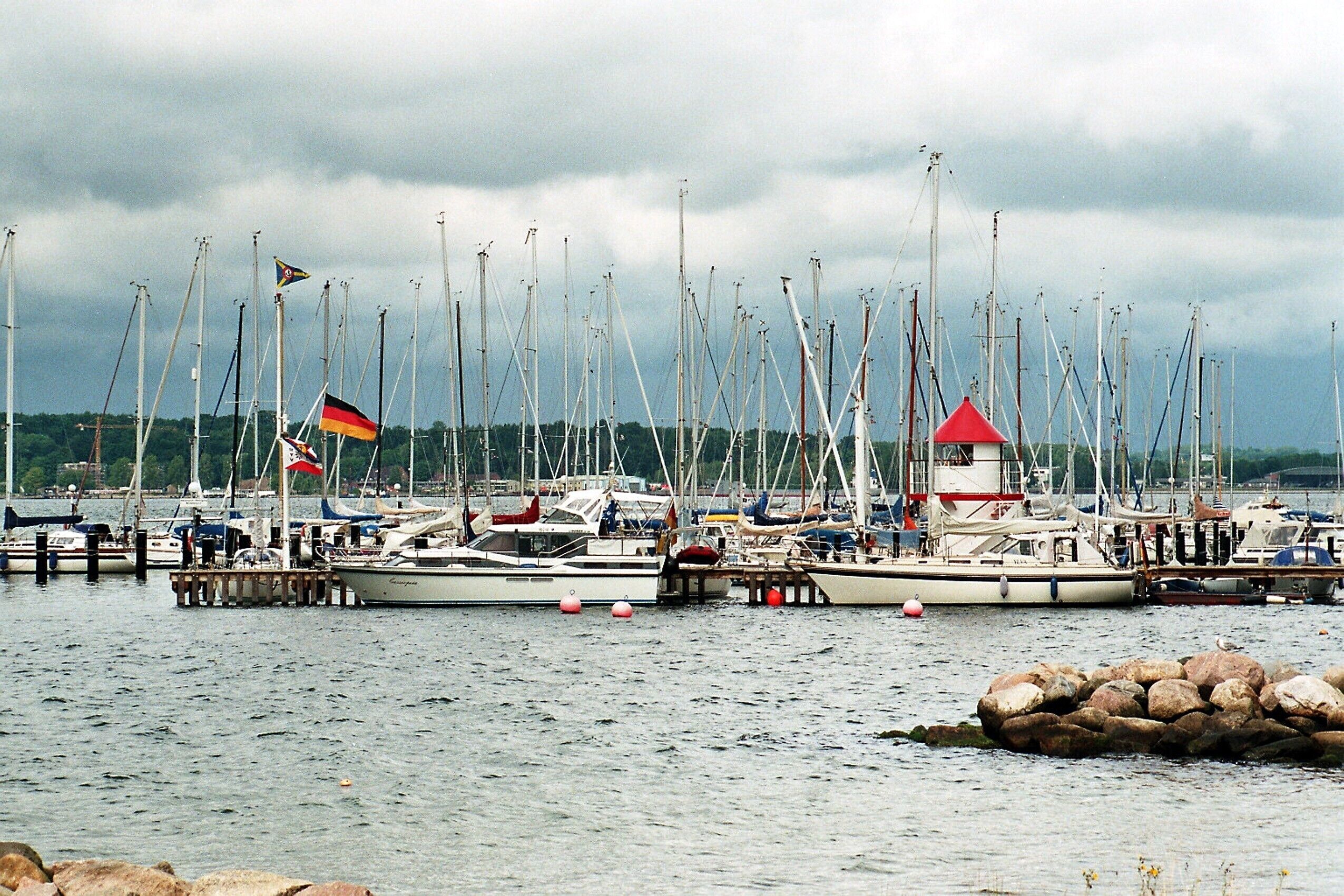 A marina with multiple sailboats docked, a lighthouse, and a German flag.