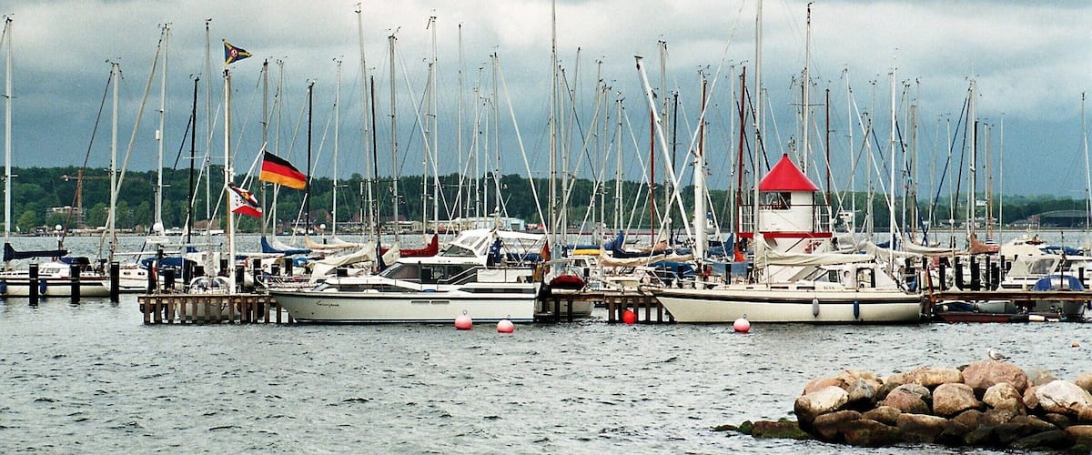 A marina with multiple sailboats docked, a lighthouse, and a German flag.