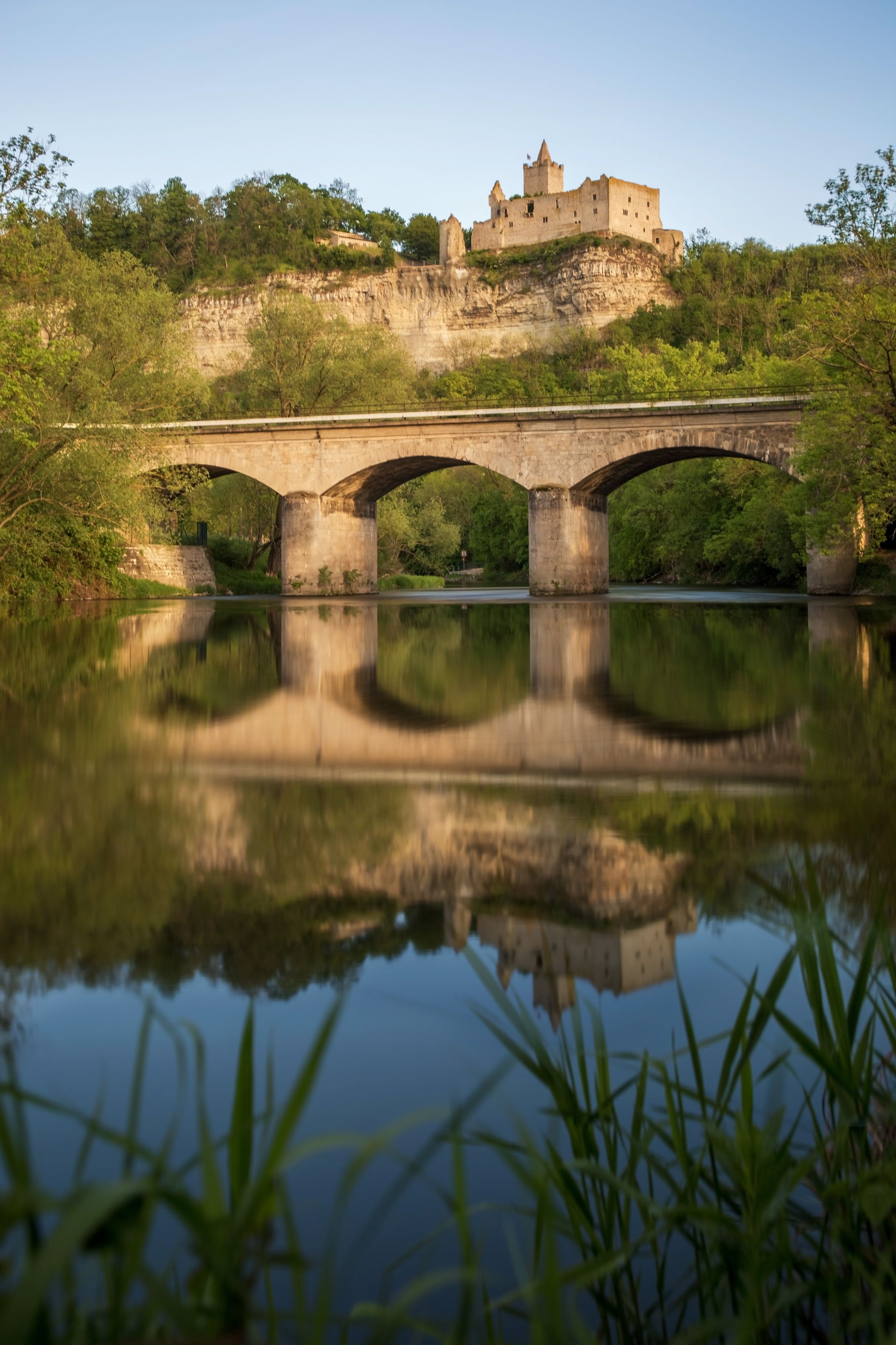 Un puente de piedra sobre un río tranquilo que refleja la vegetación circundante y un castillo en una colina al fondo.