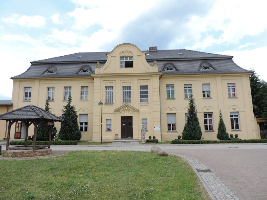A large, yellow building with a symmetrical facade, a central entrance, and a small gazebo in the foreground.