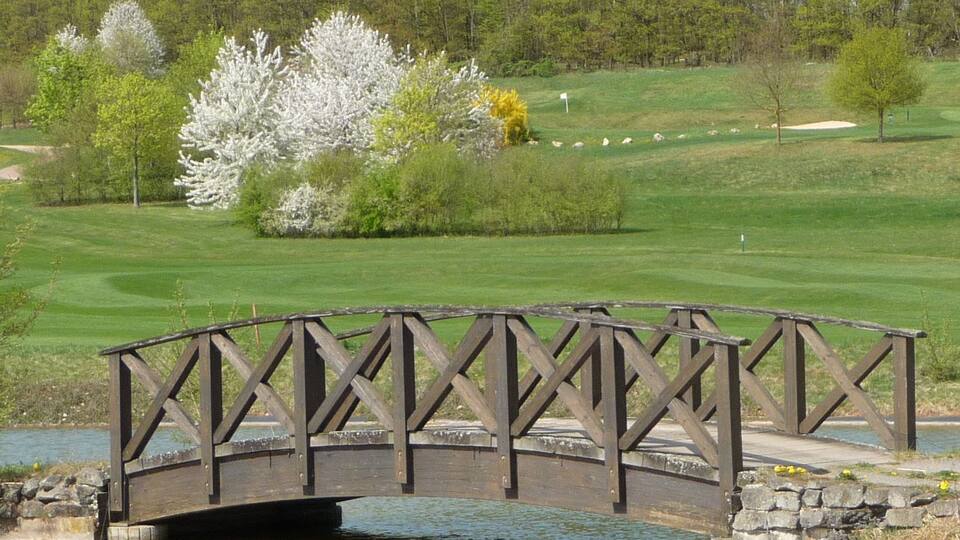 A wooden bridge over a calm body of water, surrounded by lush greenery and a golf course in the background.
