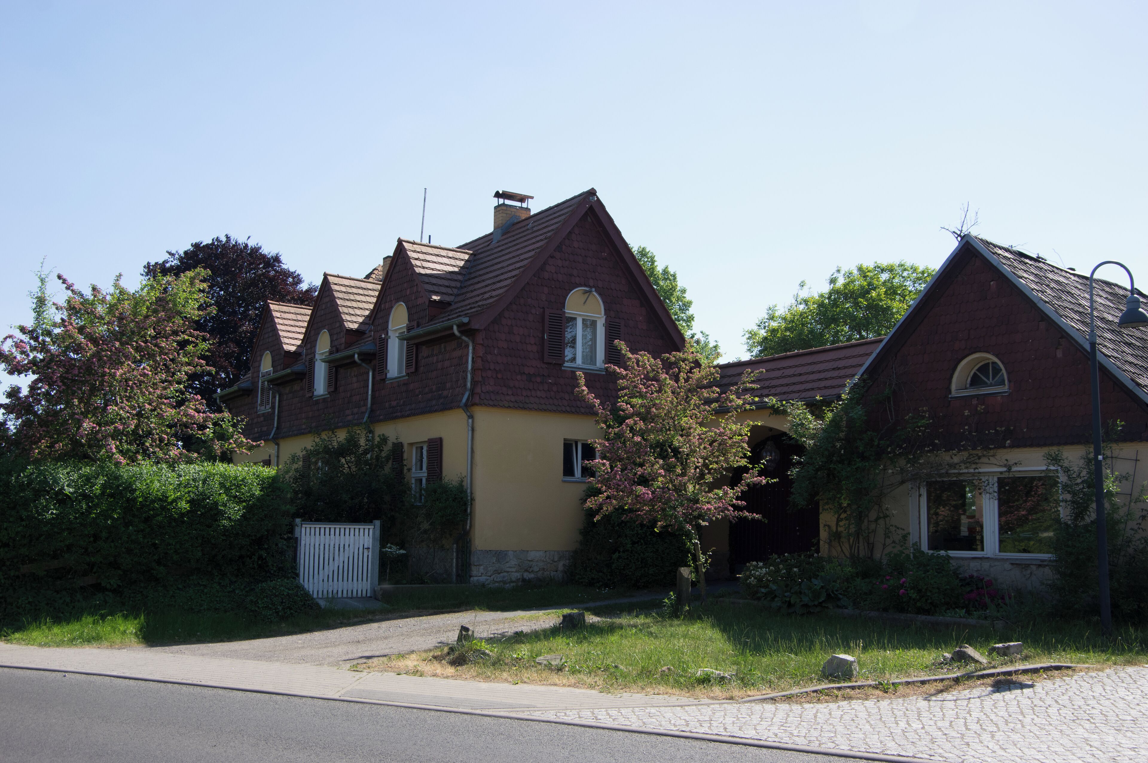 A row of two-story houses with red roofs and yellow walls, surrounded by greenery and a paved driveway.