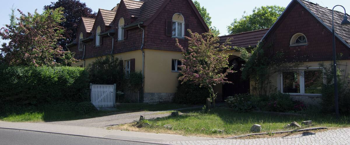 A row of two-story houses with red roofs and yellow walls, surrounded by greenery and a paved driveway.